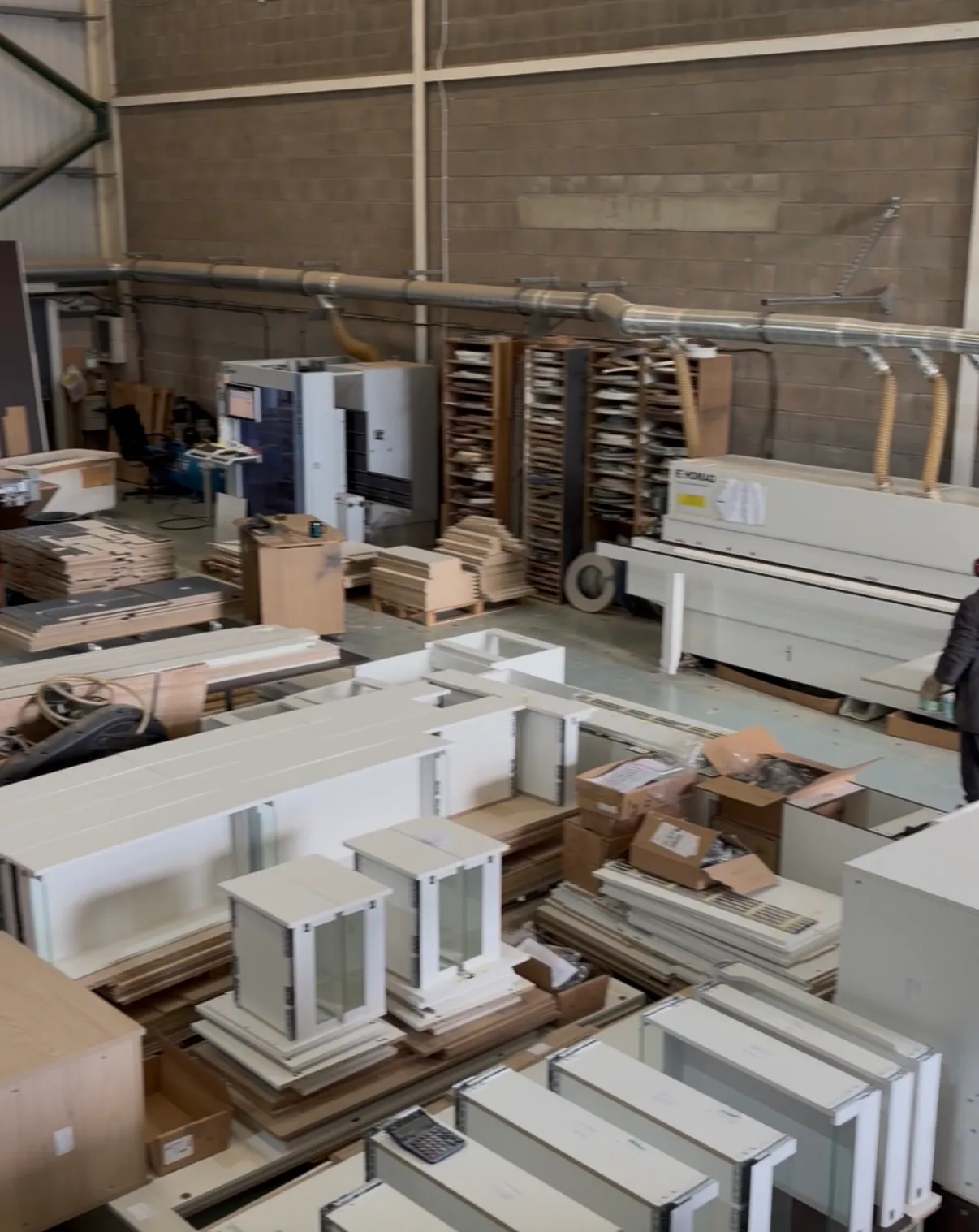 The workshop floor, with painted cabinet carcasses and stacked panels laid out in front of a beam saw and edge bander